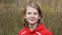 Portræt af Gustav Kalmar, dreng 14 år står og smiler, foran en grøn baggrund (hæk) med en rød hættetrøje på.